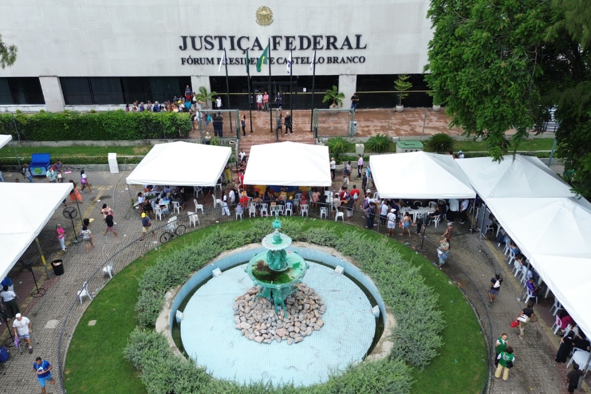Vista aérea do pátio da Justiça Federal, onde tendas brancas enfileiradas atendem diversas pessoas em um mutirão social. Ao centro, destaca-se um chafariz circular com base de pedras e uma estátua verde, cercado por gramado e arbustos baixos. Ao fundo, o imponente edifício cinza exibe o letreiro "JUSTIÇA FEDERAL - FÓRUM PRESIDENTE CASTELLO BRANCO" sob bandeiras hasteadas.